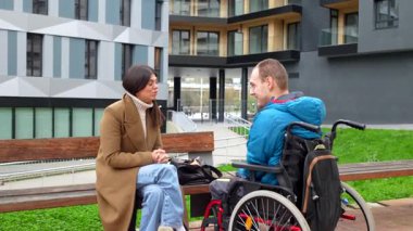 Destekleyici kadın sosyal hizmet görevlisi, tekerlekli sandalyede fiziksel engelli genç bir adamla keyifli bir konuşma yapıyor, modern bir şehir avlusunda bankta birlikte oturuyor.