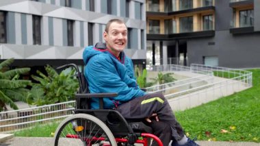 Fiziksel engelli neşeli bir gencin portresi. Tekerlekli sandalyesinde oturuyor, gülümsüyor ve gülüyor. Modern bir şehir parkında kameraya bakıyor.