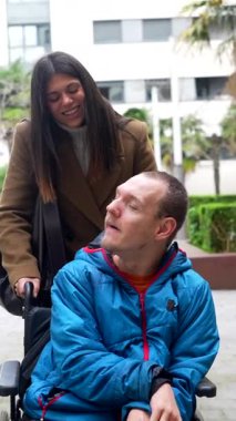 Tekerlekli sandalyedeki mutlu sakat genç adam, güzel bir bayan bakıcı ya da arkadaşla gülümsüyor ve konuşuyor. Şehir parkında birlikte yürümekten keyif alıyor.