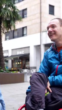 Fiziksel engelli neşeli bir adam, şehirde bir caddede tekerlekli sandalyesinde oturuyor, kamera dışında biriyle gülüyor ve konuşuyor, dayanıklılık ve pozitif bir tutum sergiliyor.