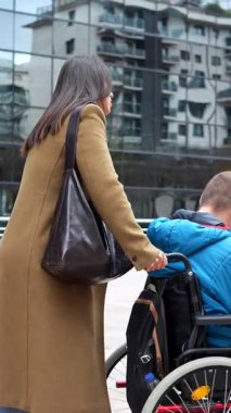 Tekerlekli sandalyedeki bir adama yardım eden kendini adamış bir kadın modern bir şehirde yol alırken, arka planda cam binalar, destek, bakım ve kentsel ulaşımı yansıtıyor.