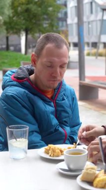 Özürlü genç beyaz bir adam dışarıda bir restoranda bakıcısıyla öğle yemeği yiyor. Asistanı, hastaya çatalla küçük lokmalar yedirerek yardımcı olur.