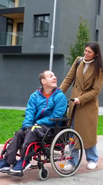 Tekerlekli sandalyede sakat bir adama yardım eden bakıcı, şehrin caddesinde yürürken konuşurken ve gülümserken, modern bir binanın yanında keyifli bir yürüyüş yaparken.