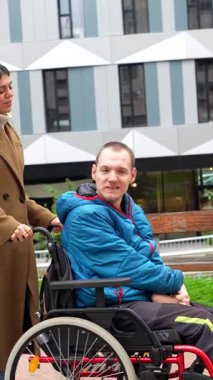 Genç bir bakıcı, tekerlekli sandalyede fiziksel engelli bir adama yardım ediyor, modern bir binanın dışındaki bir şehir parkında yürüyüş yapıyor, destek, bakım ve kapsama alanlarını temsil ediyor.