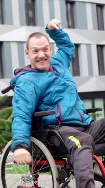 Tekerlekli sandalyede oturan, kollarını kaldıran ve modern bir şehir binasının önünde zaferi ya da başarıyı kutlayan engelli bir genç.