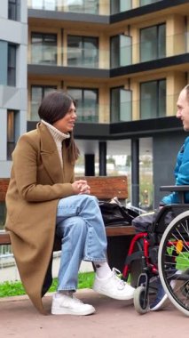 Genç bir sosyal hizmet görevlisi tekerlekli sandalyede engelli bir adamla destekleyici bir konuşma yapıyor, modern şehir parkı ortamında bir bankta birlikte oturuyor, dahil edilme ve bağlanmayı teşvik ediyor.