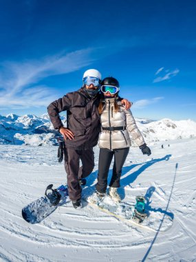 Güneşli bir dağ zirvesinde kar sörfçüleri, kasklı ve gözlüklü gülümseyen bir çift kış sporlarının, karla kaplı manzaranın, açık mavi gökyüzünün ve kaygısız maceranın tadını çıkarıyorlar.