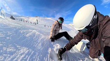 Kayak kıyafetli genç bir çift, güneşli bir günde, kalabalık bir kar yamacında nefes kesici arka plan manzarasını dolduran görkemli karlı dağlarda mola verip sohbet ediyorlar.
