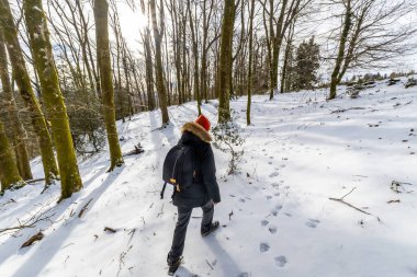 Woman walking along a snow covered path, leaving footprints in the fresh snow while exploring a tranquil winter forest with bare trees and bright sunlight