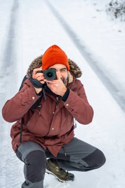 Turuncu bir bere takan fotoğrafçı ve parka karda diz çöküp, soğuk bir kış gününde doğal bir ortamda dijital bir kamerayla görüntüleri çekiyor.