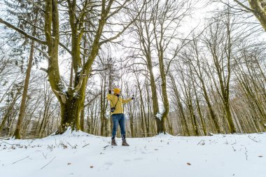Sarı ceketli ve bereli bir adam kış yürüyüşünün tadını çıkarıyor, karla kaplı bir ormanda dikiliyor, uzun çıplak ağaçlarla çevrili, yukarı bakıyor ve kollarına uzanıyor.