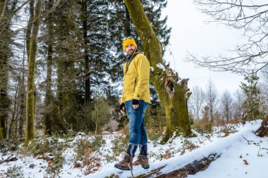 Sarı ceketli ve bereli bir adam karlı bir ormanda düşmüş bir ağacın gövdesinde duruyor, doğanın tadını çıkarıyor ve yürüyüş sırasında soğuk havanın tadını çıkarıyor.