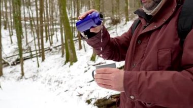 Sırt çantalı orta yaşlı bir adam soğuk bir kış günü güzel bir karlı ormanda yürüyüp doğanın tadını çıkarırken bir şişeden ılık bir içecek doldurmak ve içmek için mola veriyor.