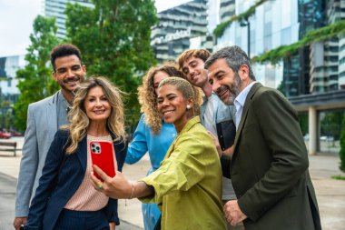 Çeşitli güler yüzlü iş adamları ve meslektaşları modern binalarla dolu bir şehir merkezinde grup halinde selfie çekiyorlar.