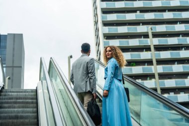 Two diverse business colleagues ascending an escalator, looking towards a modern city building, symbolizing career growth, collaboration, and urban professional life with focus and determination