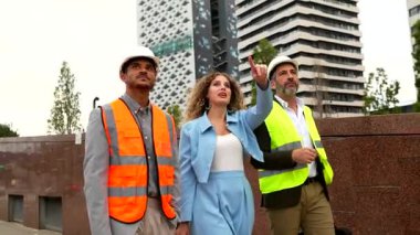 Mimarlar ve mühendislerden oluşan çeşitli bir ekip bir inşaat sahasında yürürken, projenin ilerlemesini tartışırken arka plandaki modern gökdeleni işaret ediyor ve planları inceliyor.