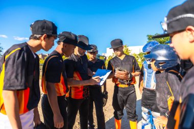 Siyah ve turuncu üniformalı beyzbol koçu, açık mavi bir gökyüzünün altında, sahada formalı gençlerle dolu bir panoda stratejiyi gözden geçirdi.