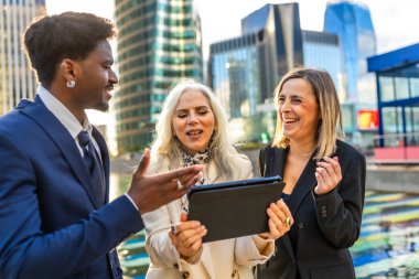Çeşitli iş adamları dışarıda canlı bir toplantı yapıyor, dijital tablete bakıyor ve modern bir şehir manzarasıyla çevrili yeni bir projede işbirliği yapıyorlar.