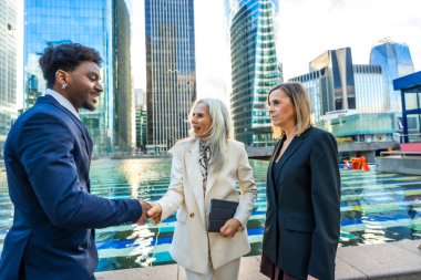 Çeşitli iş adamları, siyahi bir adam ve iki kadın, el sıkışıyor ve modern bir finans bölgesinde cam gökdelenlerle ışığı yansıtan tartışmalar yapıyorlar.