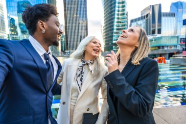 Şehirdeki açık havada ayakta durup, çeşitli iş arkadaşları birlikte gülerken, arka planda kentsel binaların olduğu kurumsal bir aranın keyfini çıkarıyorlar.