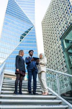 Şehir merkezindeki bir şirket bölgesinde çeşitli iş adamları toplanıyor, arkasında gökdelenler olan bir tablet üzerinde işbirliği yapıyorlar, takım çalışmasını ve kentsel yeniliği sembolize ediyorlar.