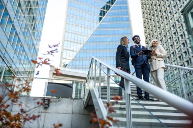 Çeşitli iş adamları gülümseyip etkileşime girerken modern açık hava merdivenlerinde kurumsal binalarla birlikte bir tablete bakıyorlar, proje fikirleri ve stratejilerini tartışıyorlar.