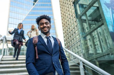 Afrika kökenli Amerikalı iş adamı kameraya gülümsüyor ve modern bir şehir ortamında merdivenlerden çıkıyor. Kadın meslektaşları arka planda yükseliyor. Başarı ve kariyer büyümesini aktarıyor.