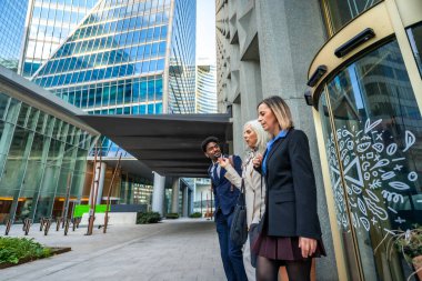 Çağdaş ofis binaları ve döner kapılı modern bir şehir ortamında gezinirken, çeşitli iş arkadaşları birlikte yürüyor ve konuşuyor, fikirleri paylaşıyor.