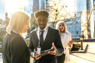 İş arkadaşları açık hava kentsel toplantılarında iş birliği yapıyor, dijital tablet üzerindeki bilgileri incelerken fikir alışverişinde bulunuyorlar ve arka planda cep telefonuyla konuşan bir kadın iş arkadaşı da var.