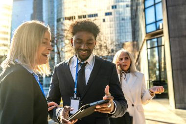 Şehirde birlikte çalışan çeşitli iş adamları tablete bakarken fikir alışverişinde bulunuyorlar ve gülümsüyorlar. Arka planda telefonla konuşan başka bir meslektaşları var.