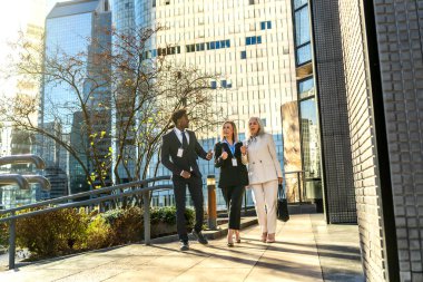 Üç farklı iş adamı, bir siyah adam ve iki beyaz kadın, güneşli bir günde modern finans bölgesinde yürüyorlar, tartışıyorlar.