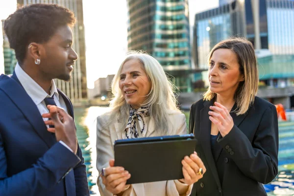 Modern bir şehir ortamında yapılan bir açık hava toplantısında dijital tablet kullanarak iş birliği yapan ve yeni stratejileri tartışan çok ırklı iş adamları grubu.