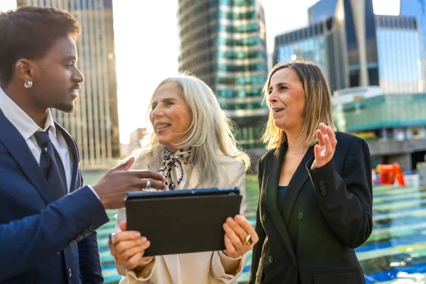 Çeşitli iş adamları, arka planda modern şehir binalarının bulunduğu dijital tablete bakarken bir projeyi tartışarak gayri resmi bir açık hava toplantısına giriyorlar.