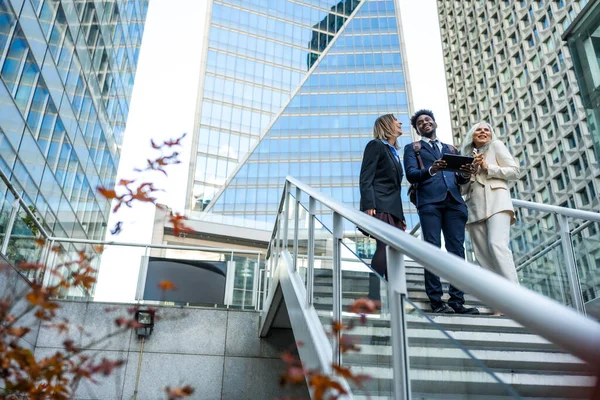 Çeşitli iş adamları gülümseyip etkileşime girerken modern açık hava merdivenlerinde kurumsal binalarla birlikte bir tablete bakıyorlar, proje fikirleri ve stratejilerini tartışıyorlar.
