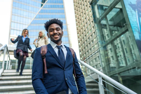 Afrika kökenli Amerikalı iş adamı kameraya gülümsüyor ve modern bir şehir ortamında merdivenlerden çıkıyor. Kadın meslektaşları arka planda yükseliyor. Başarı ve kariyer büyümesini aktarıyor.