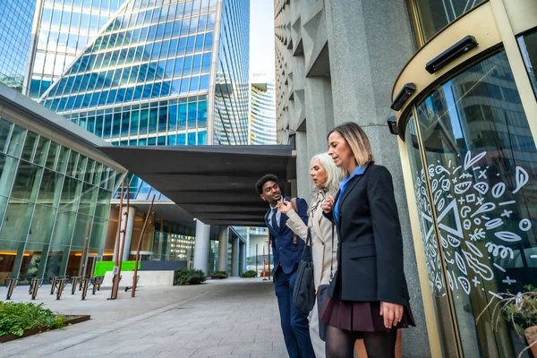 Çağdaş ofis binaları ve döner kapılı modern bir şehir ortamında gezinirken, çeşitli iş arkadaşları birlikte yürüyor ve konuşuyor, fikirleri paylaşıyor.