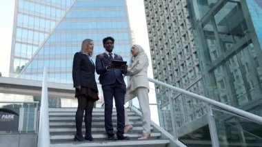 Mesleki çeşitlilikteki iş arkadaşları, bir tablet üzerinde projeyi tartışırken modern bir şehirde, merdivenlerde dikilirken ve arka planda cam gökdelenler varken kurumsal bir toplantı sırasında