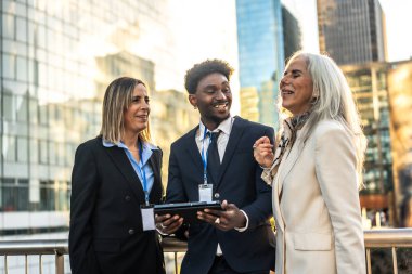 Modern kentsel bir ortamda yapılan bir açık şehir toplantısında dijital tablet hakkındaki bilgileri inceleyen, takım çalışmasını teşvik eden ve işbirliği yapan çeşitli iş adamları