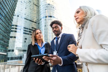 Dijital tablet üzerinde işbirliği yapan, modern cam gökdelenlerin önünde duran, şehir hayatını, takım çalışmasını ve küresel iş stratejilerini sembolize eden çeşitli iş adamları