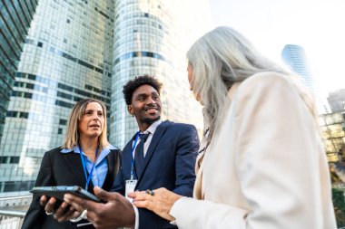 Modern bir finans bölgesinde düzenlenen bir açık hava toplantısında proje ve fikirleri tartışan çeşitli iş adamları, takım çalışması, strateji ve küresel iş dünyası kavramlarını yansıtıyorlar.