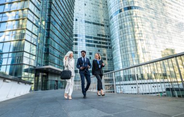 Modern bir finans bölgesinde cam ofis binaları parlak gökyüzünü yansıtırken çeşitli iş adamları yürür, iletişim kurar ve güler.