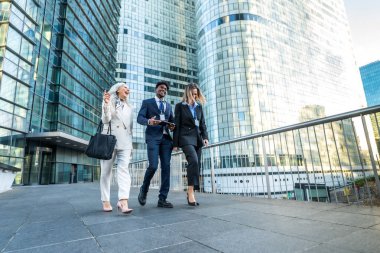 Aralarında kıdemli bir kadının da bulunduğu çeşitli iş adamları, şehir geçidinde birlikte yürüyorlar, ellerinde belgelerle arka planda modern cam gökdelenlerle iş konuşuyorlar.