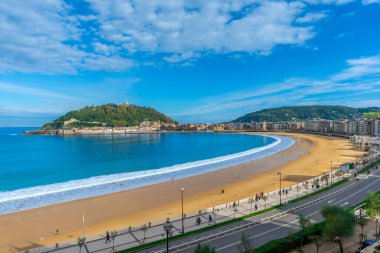 San Sebastian körfezi boyunca La concha plajı kıvrılıyor. Kum ve gezinti güvertesinde yürüyen insanlar, Monte igueldo 'ya ve şehir sahillerine tepeden bakan parlak mavi bir gökyüzü altında.