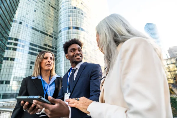 Modern bir finans bölgesinde düzenlenen bir açık hava toplantısında proje ve fikirleri tartışan çeşitli iş adamları, takım çalışması, strateji ve küresel iş dünyası kavramlarını yansıtıyorlar.