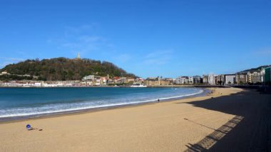 San Sebastian, İspanya 'daki ikonik La Concha sahilinin güzel panoramik manzarası kumlu körfezi, Cantabrian Denizi' nin sakin dalgalarını ve açık mavi gökyüzünün altındaki şehir siluetini gösteriyor.