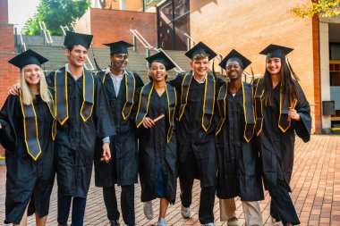 Üniversite mezuniyetlerini kutlayan bir grup genç öğrenci kampüste birlikte yürürken gururla kep, cübbe ve çalgılar giyiyorlar.