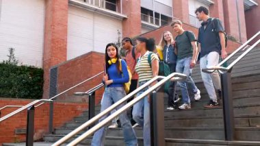 Sırt çantaları ve not defterleriyle kampüs binasının merdivenlerinden aşağı inen ve derslerini bitirdikten sonra gülümseyen çok ırklı genç üniversite öğrencileri grubu.