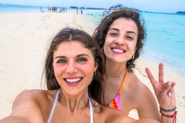 Gülen kadınlar, güzel beyaz bir kumsalda turkuaz suyla neşeli bir selfie çekiyorlar. Maldivler 'e tropikal bir tatil sırasında, dostluğu ve seyahat anılarını sembolize ediyorlar.