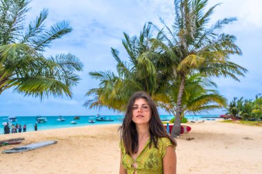 Maldivler içinde tropik bir plajda duran bir kadın. Etrafı kum, palmiye ağaçları ve turkuaz okyanus suyuyla çevrili. Arka planda bulutlu bir gökyüzünün altında görünen tekneler var.