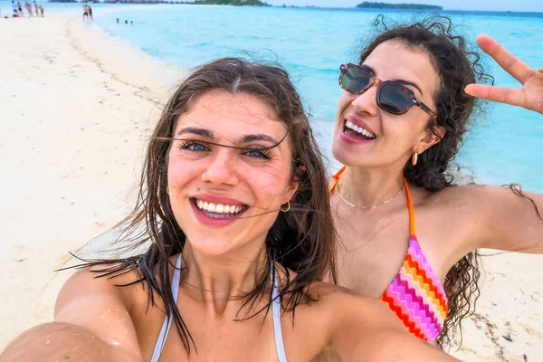 İki gülümseyen genç kadın, arka planda turkuaz okyanusla beyaz kumlu bir plajda kendi portresini çekiyorlar. Tropikal tatillerinde neşe ve dostluklarını paylaşıyorlar.